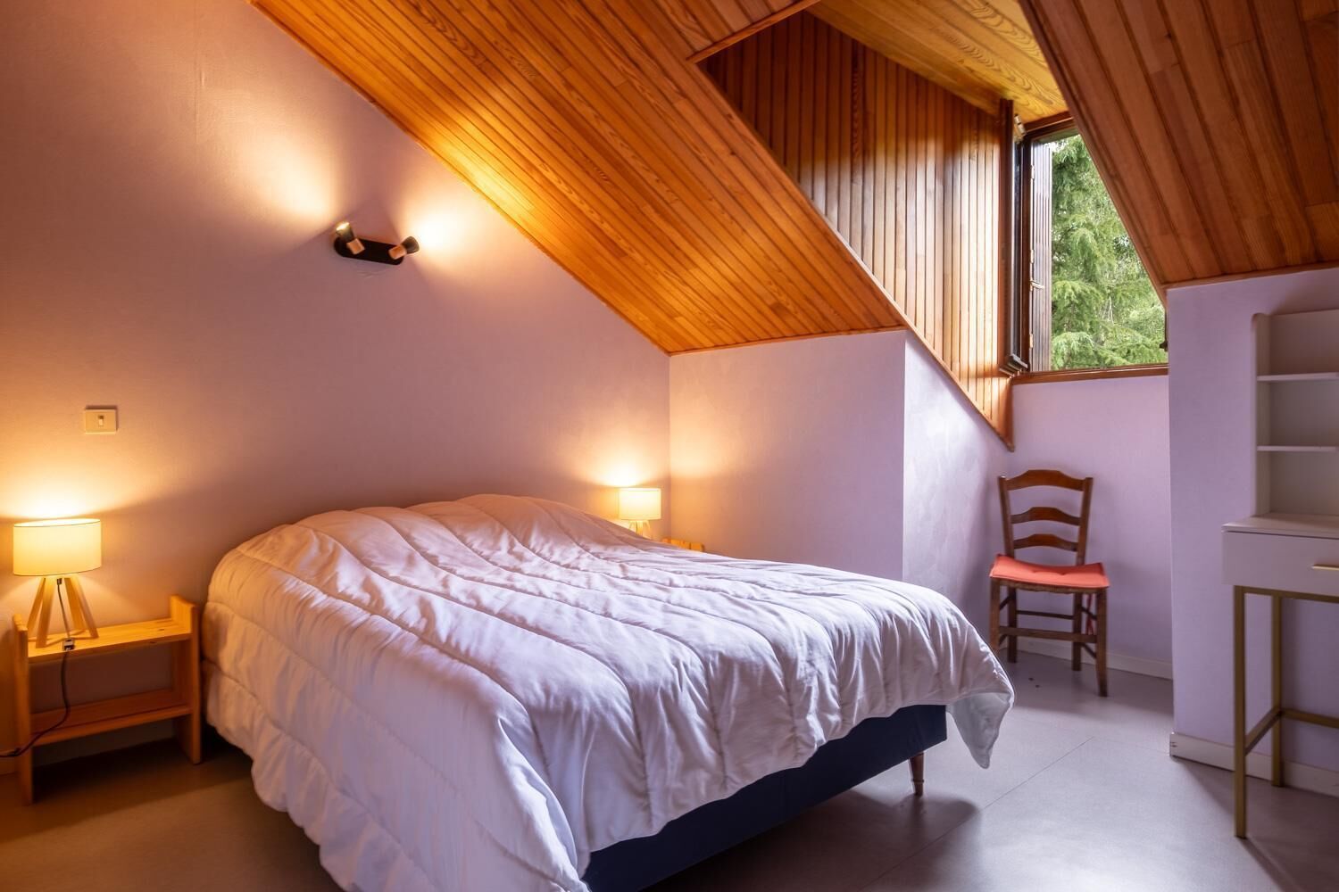 A bedroom with a sloped ceiling, a bed, a chair, a desk, and a window.