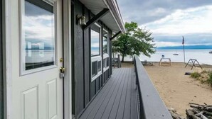 Eine Holzterrasse mit einer weißen Tür, großen Fenstern und Blick auf einen Strand mit einer Flagge.