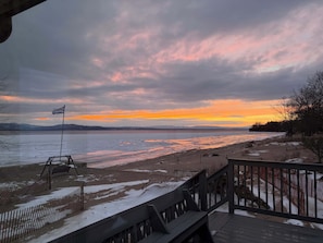 A wooden deck overlooking a beach with a flag, a view of the water, and distant mountains. - The Lake House – Lake Champlain Waterfront Retreat (Colchester)