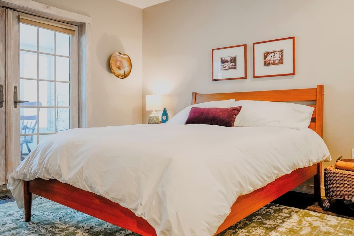 A hotel room with a bed, a nightstand, a lamp, two framed pictures on the wall, and a door leading to an outdoor area.
