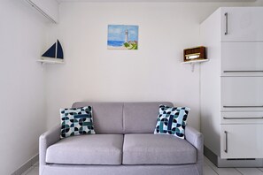 A modern living room with a grey sofa, two patterned cushions, a small sailboat shelf, a lighthouse painting, and a radio on a shelf.