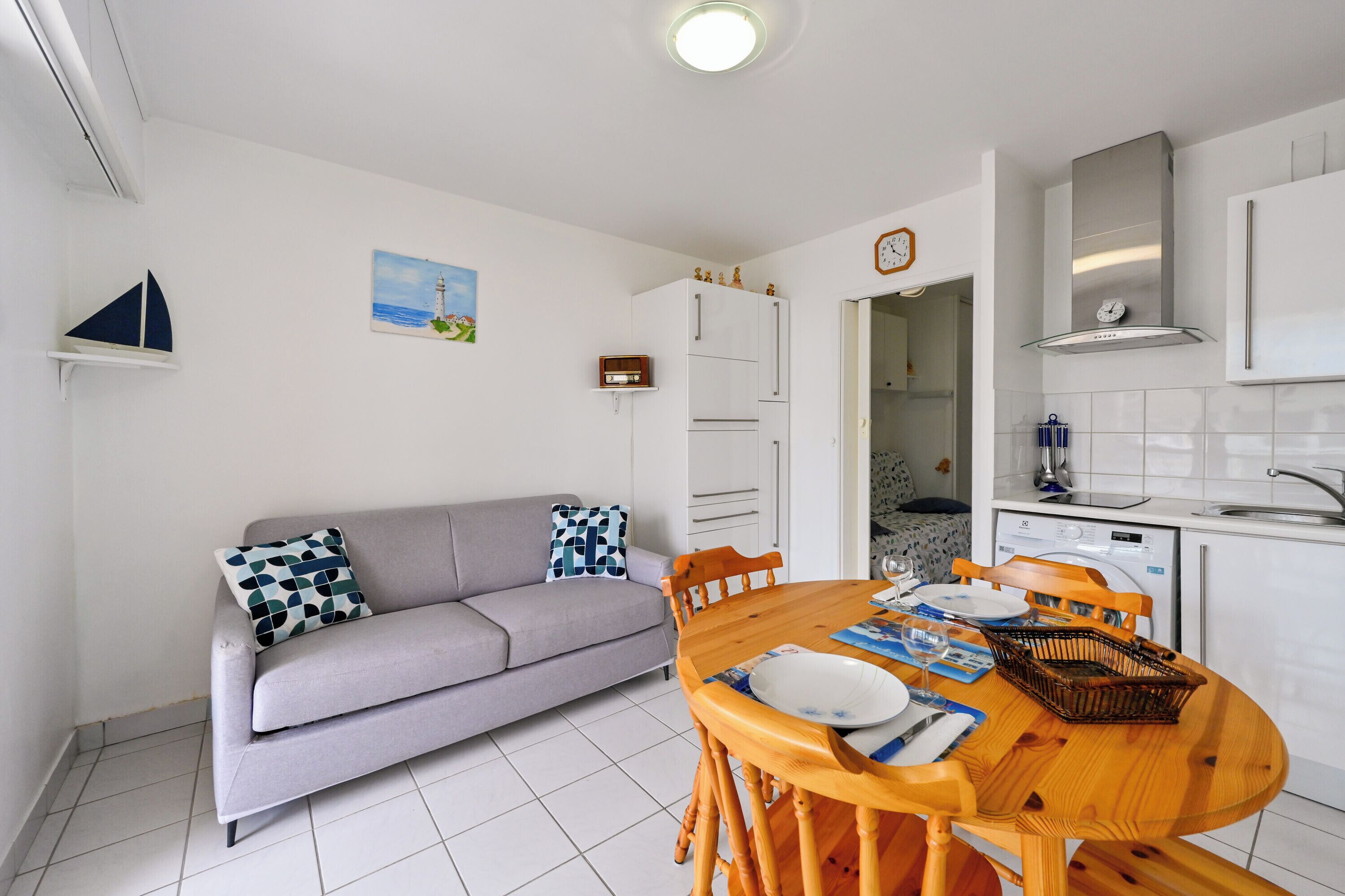 Een gezellig, compacte leefruimte met een kitchenette, een eethoek met een houten tafel en stoelen, een grijze bank met decoratieve kussens, en een kleine decoratie van een zeilboot aan de muur.