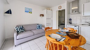 Un espacio habitable compacto con kitchenette, un comedor con mesa y sillas de madera, un sofá gris con cojines estampados y una pequeña decoración de velero en la pared.