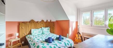 A bedroom with a sloped ceiling, a bed with a cactus-patterned bedspread, a wooden headboard, a standing lamp, a guitar, and a window with a view of greenery.