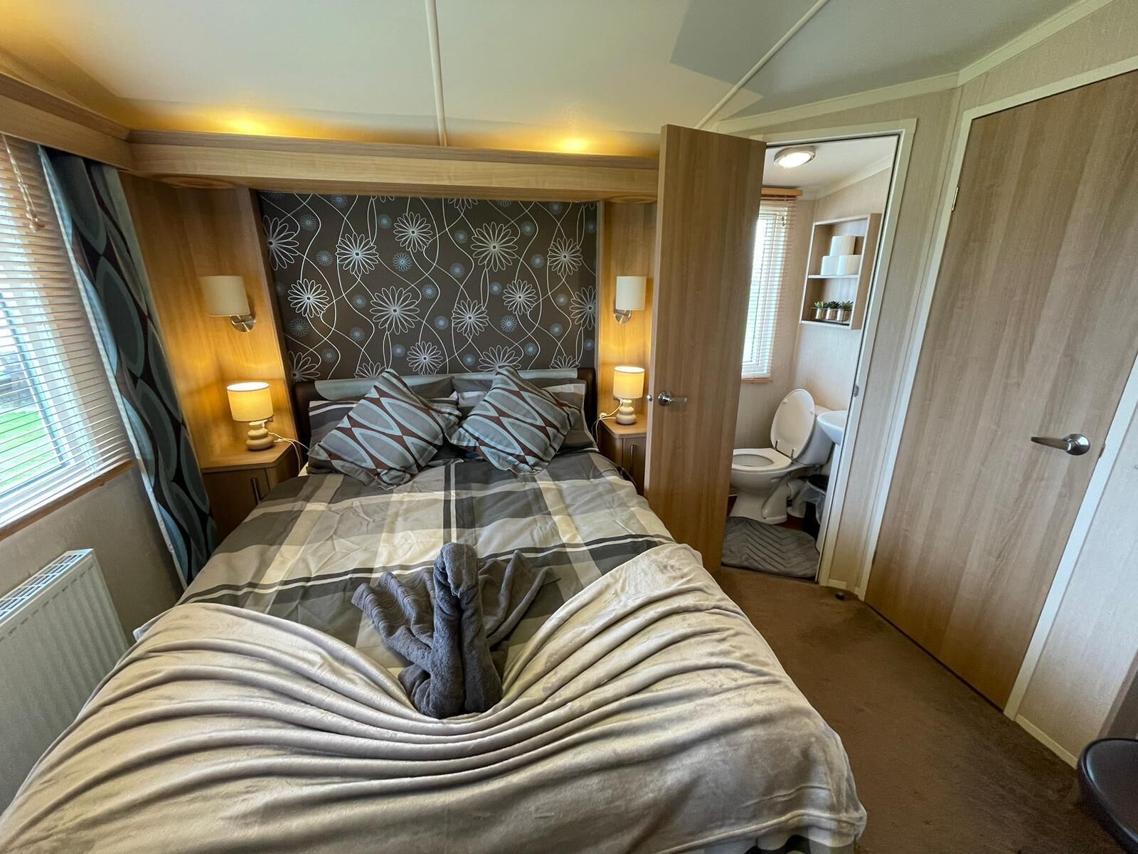 Ein Schlafzimmer mit Bett, Nachttischlampen, einem Kopfteil mit Blumenmuster, einem Badezimmer mit Toilette und Waschbecken sowie einer Tür zu einem weiteren Raum.