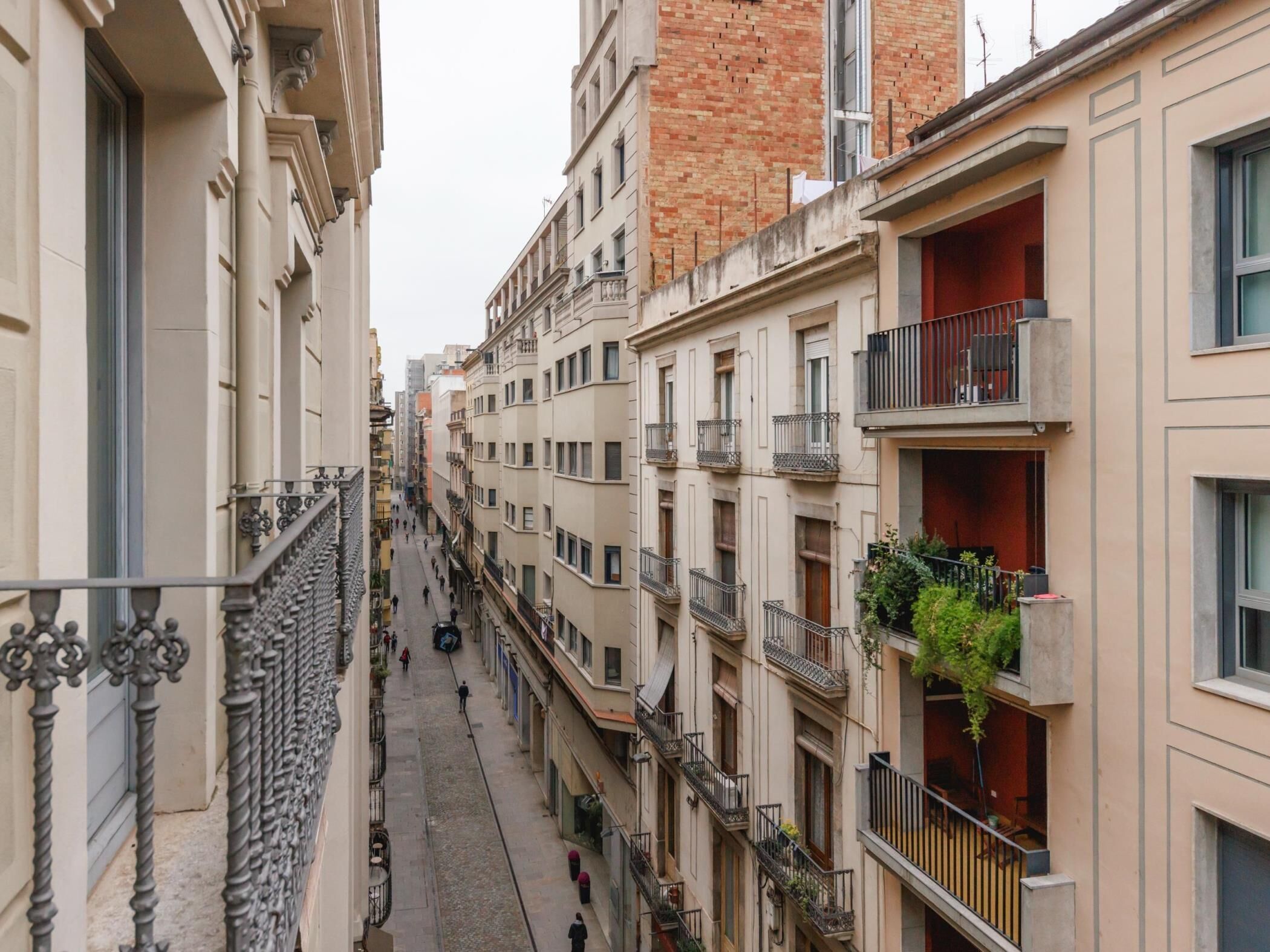 Eine schmale Stadtstraße, gesäumt von mehrstöckigen Gebäuden mit Balkonen und Geländern, sowie vereinzelten Grünpflanzen.