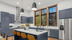 Een moderne keuken met donkere kasten, een centraal eiland met barstoelen, een roestvrijstalen koelkast en een groot raam met uitzicht op bomen.