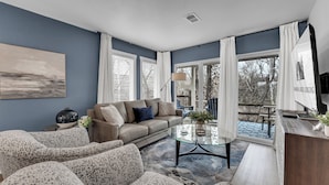 Ein modernes Wohnzimmer mit einer Couch, Sesseln, einem Glastisch und Blick ins Freie durch große Fenster.