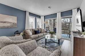 A modern living room with a sofa, armchairs, a glass coffee table, and a view of the outdoors through large windows.