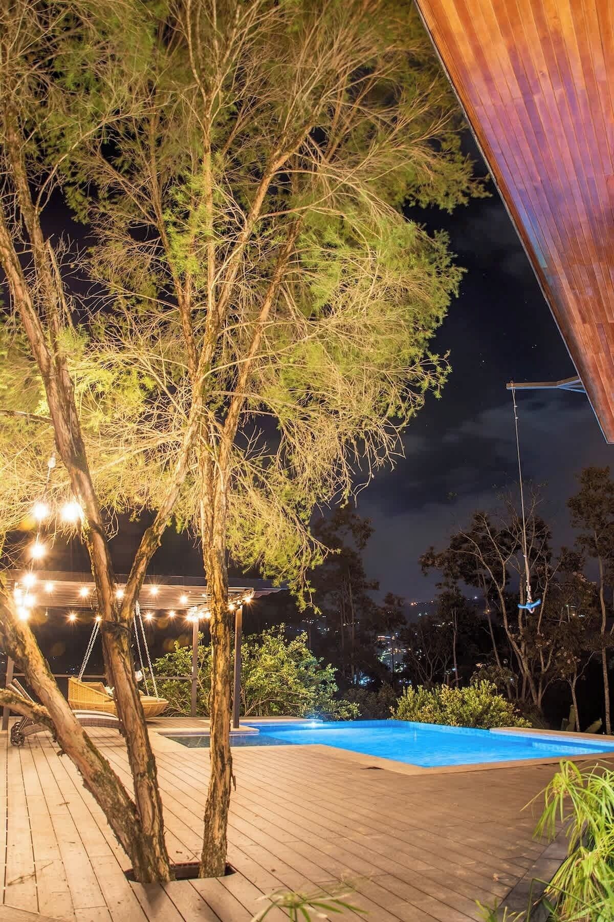A wooden deck with a swimming pool, surrounded by trees and illuminated by ambient lighting.
