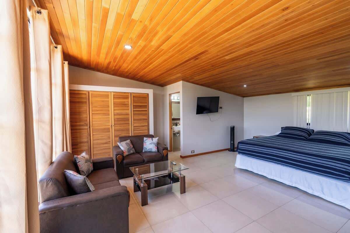 A modern living room with a wooden ceiling, a sofa, a glass coffee table, a bed, and a TV.