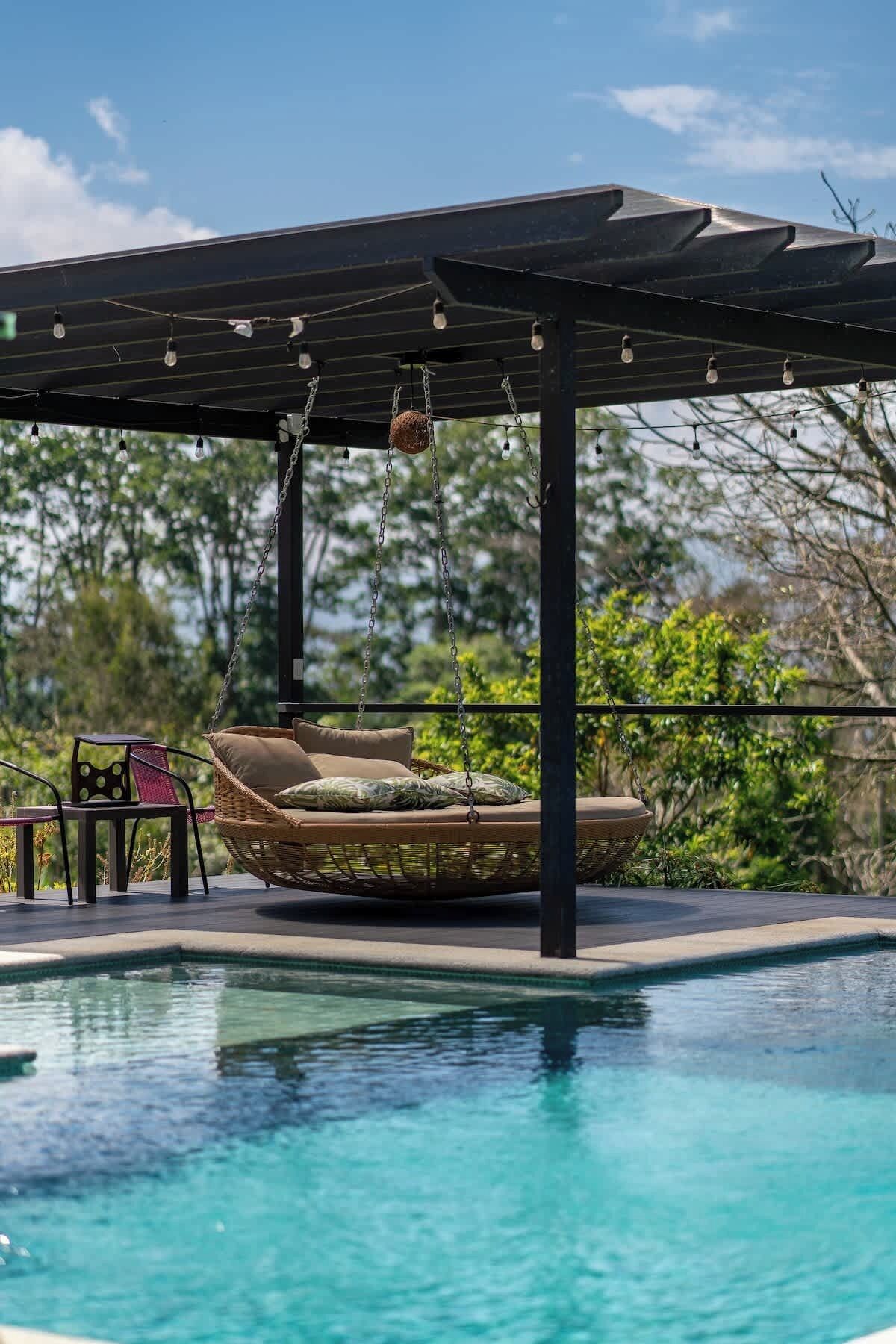 Un espace aménagé au bord de la piscine, comprenant un fauteuil suspendu, une table et une pergola illuminée de guirlandes lumineuses.