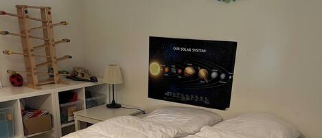 En pænt redt seng med hvide sengetæpper, et plakat af solsystemet på væggen og en træstige med legetøj på en hylde.