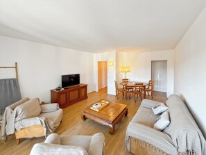Een woonkamer met een televisie, bank, salontafel en een eethoek met stoelen en een lamp.