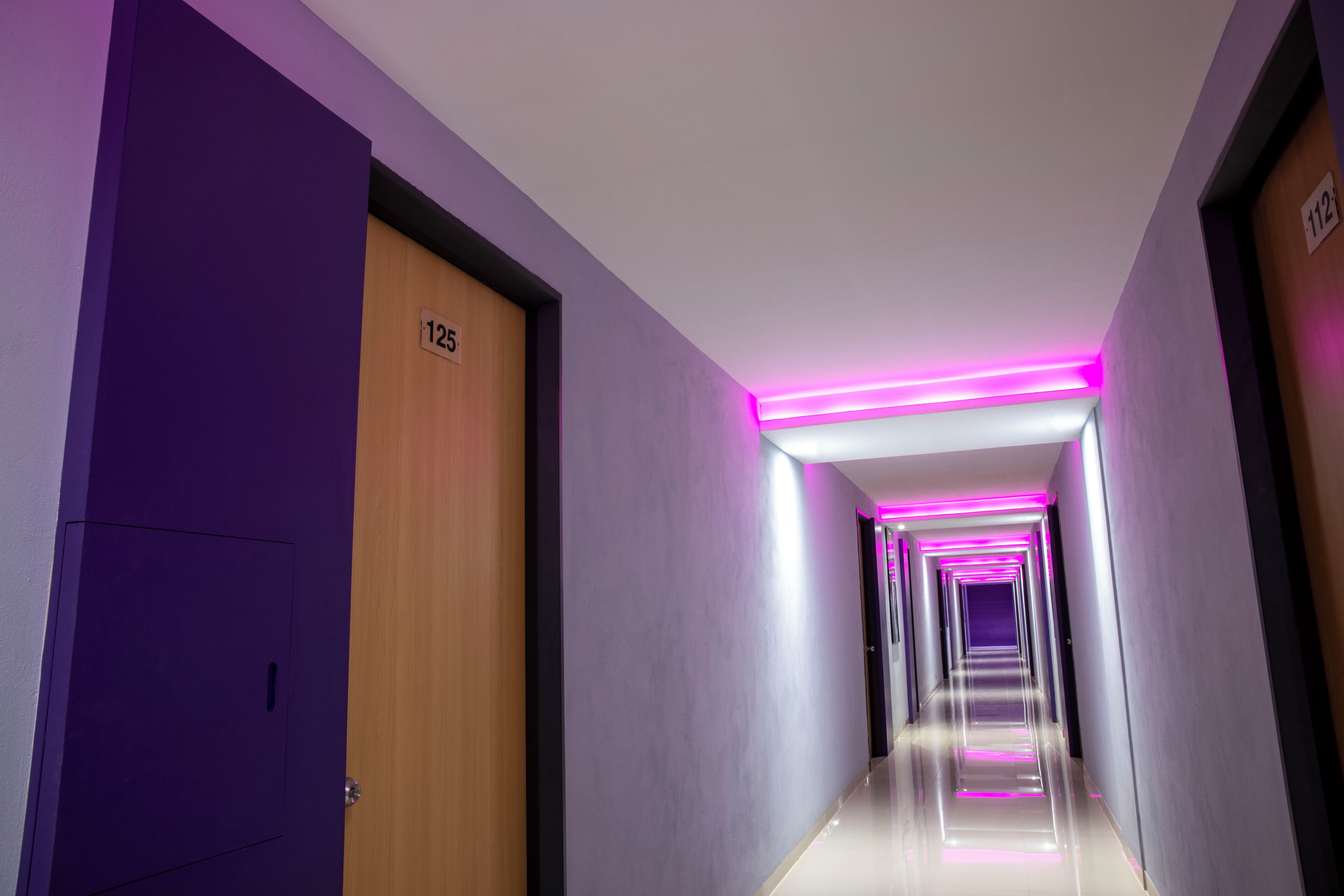 Un pasillo con paredes moradas, una puerta de madera con la etiqueta '125' y un corredor con suelo reflectante e iluminación rosada.