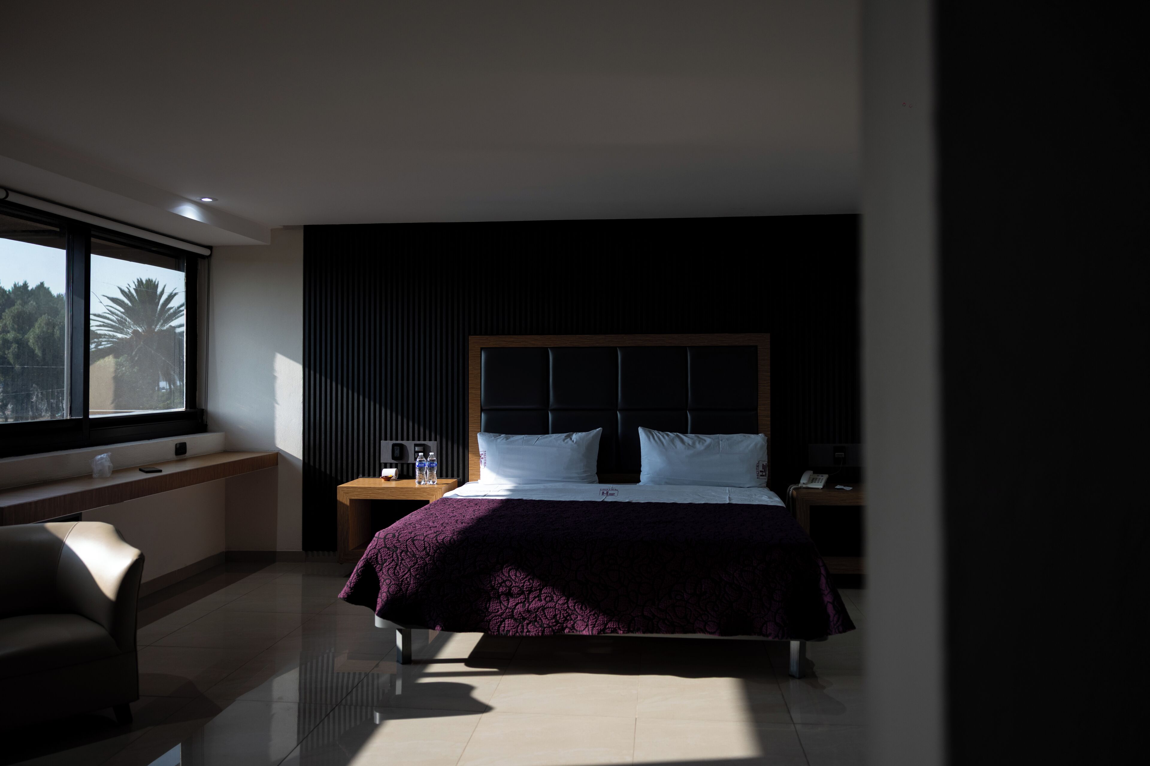 A modern hotel room with a large bed, a nightstand, a chair, and a window with a view of palm trees.