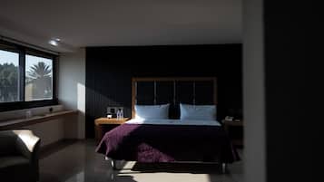A modern hotel room with a large bed, a nightstand, a chair, and a window with a view of palm trees.