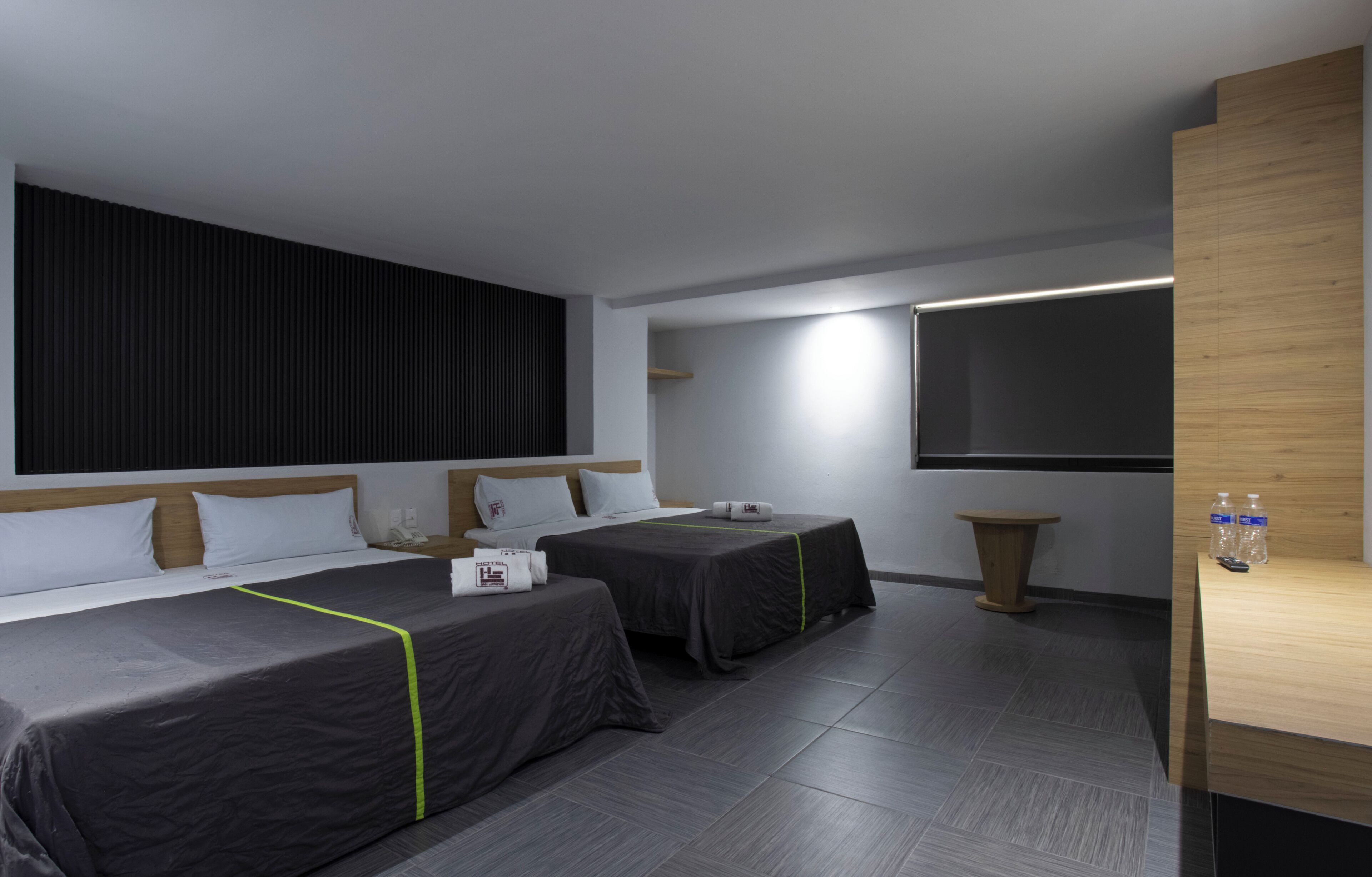 Una habitación de hotel moderna con dos camas, un televisor de pantalla plana, una pequeña mesa de madera y una botella de agua en la mesita de noche.