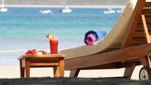 Ein Holz-Loungesessel mit Kissen, ein kleiner Tisch mit einem Getränk und einer Schleife, und im Hintergrund Blick auf Strand und Boote.