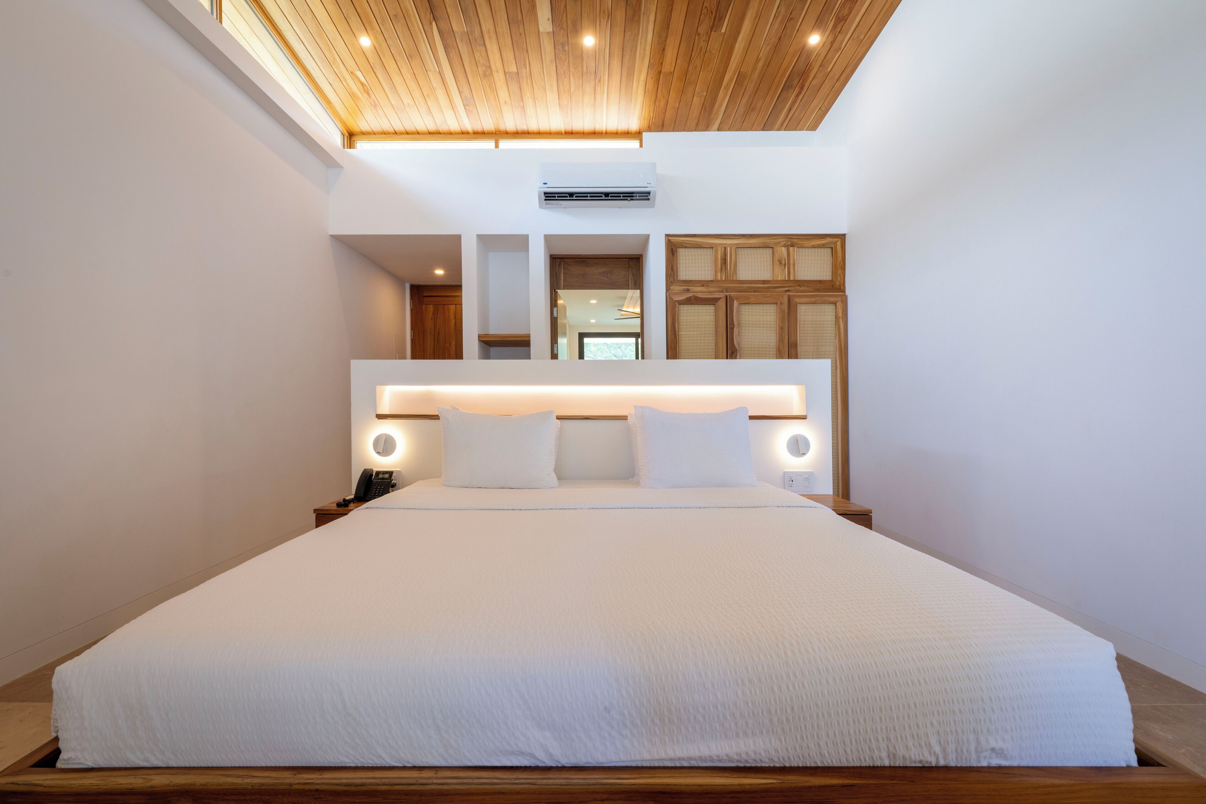 A modern hotel room with a large bed, wooden ceiling, and a built-in headboard with integrated lighting.
