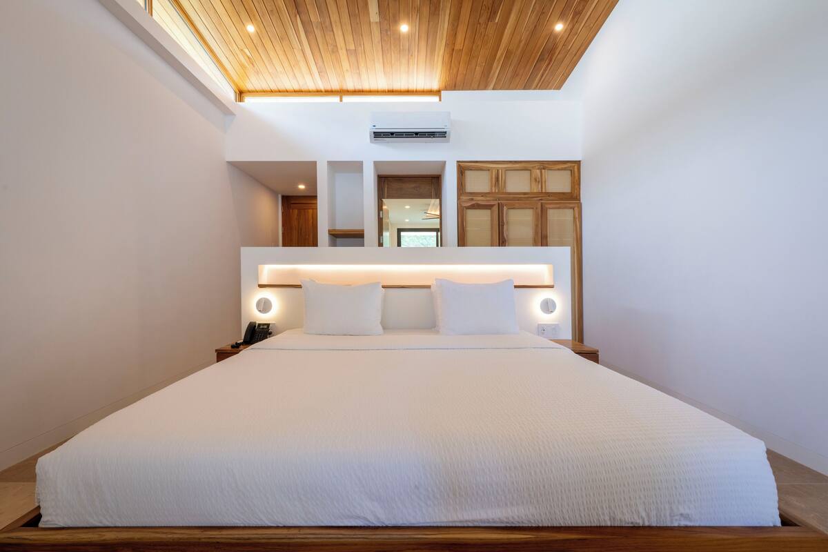 Una habitación de hotel moderna con una cama grande, techo de madera y cabecera integrada con iluminación.