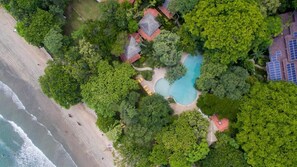 Eine Vogelperspektive zeigt einen Ferienort mit Schwimmbad, umgeben von üppiger Vegetation, Zugang zum Strand und Solaranlagen.
