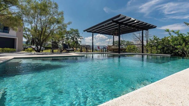 En klar swimmingpool med et pergola-liggende tagområde, omgivet af grønt og en moderne bygning.