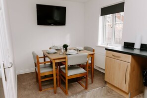 A dining area with a wooden table set for four, a black flat-screen TV mounted on the wall, and a window with a black blind.