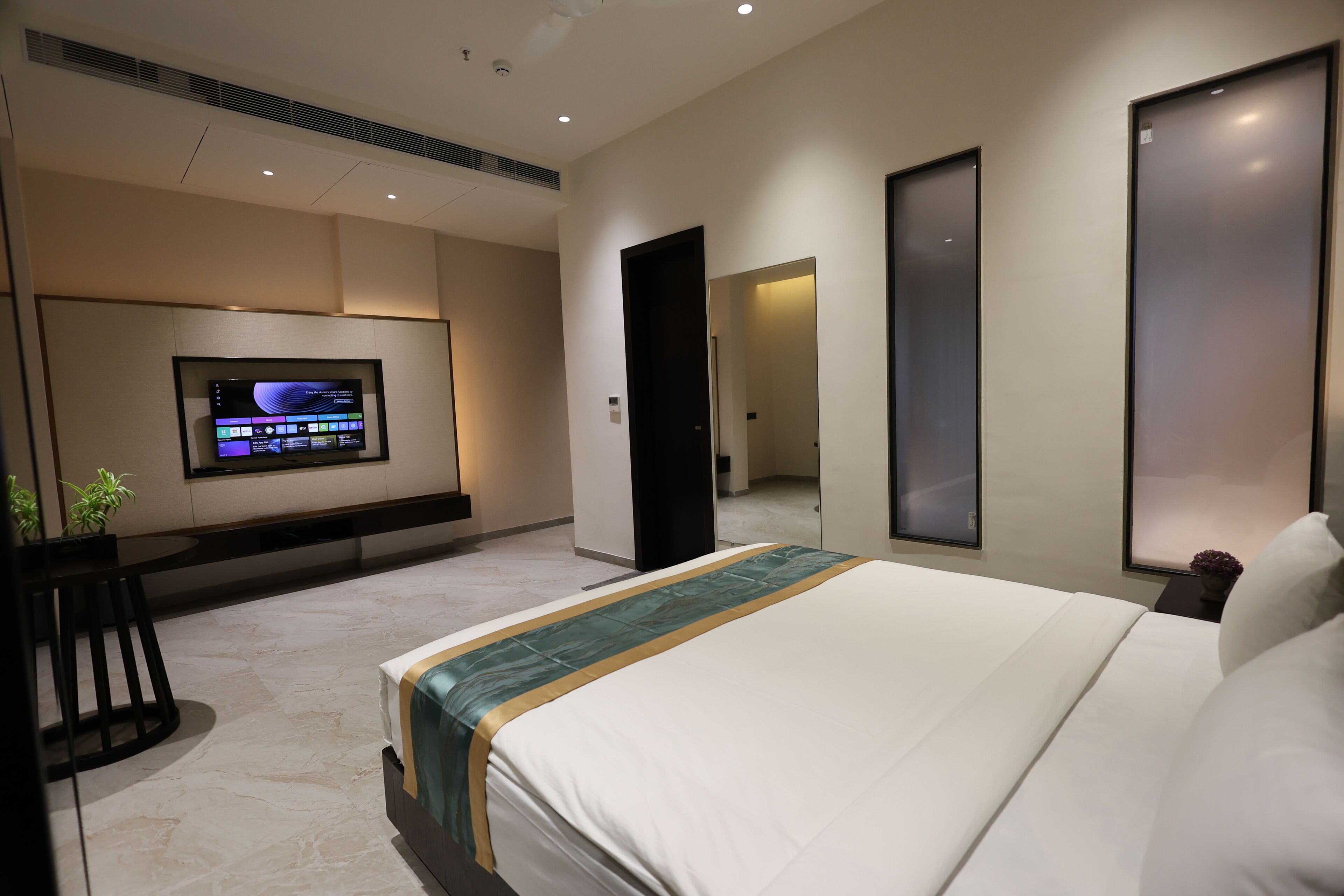A modern hotel room with a large bed, a flat-screen TV mounted on the wall, a small table with a plant, and a door leading to another room.