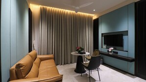 A modern living room with a sofa, a dining table with chairs, a TV mounted on the wall, and a ceiling fan. - ASHIRWAD CLUB RESORT (Jamnagar)
