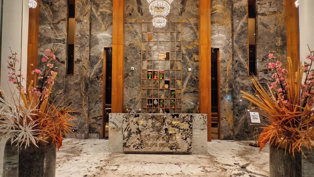 A marble reception desk with a chandelier above, wooden paneling, and a display shelf with decorative items. - ASHIRWAD CLUB RESORT (Jamnagar)