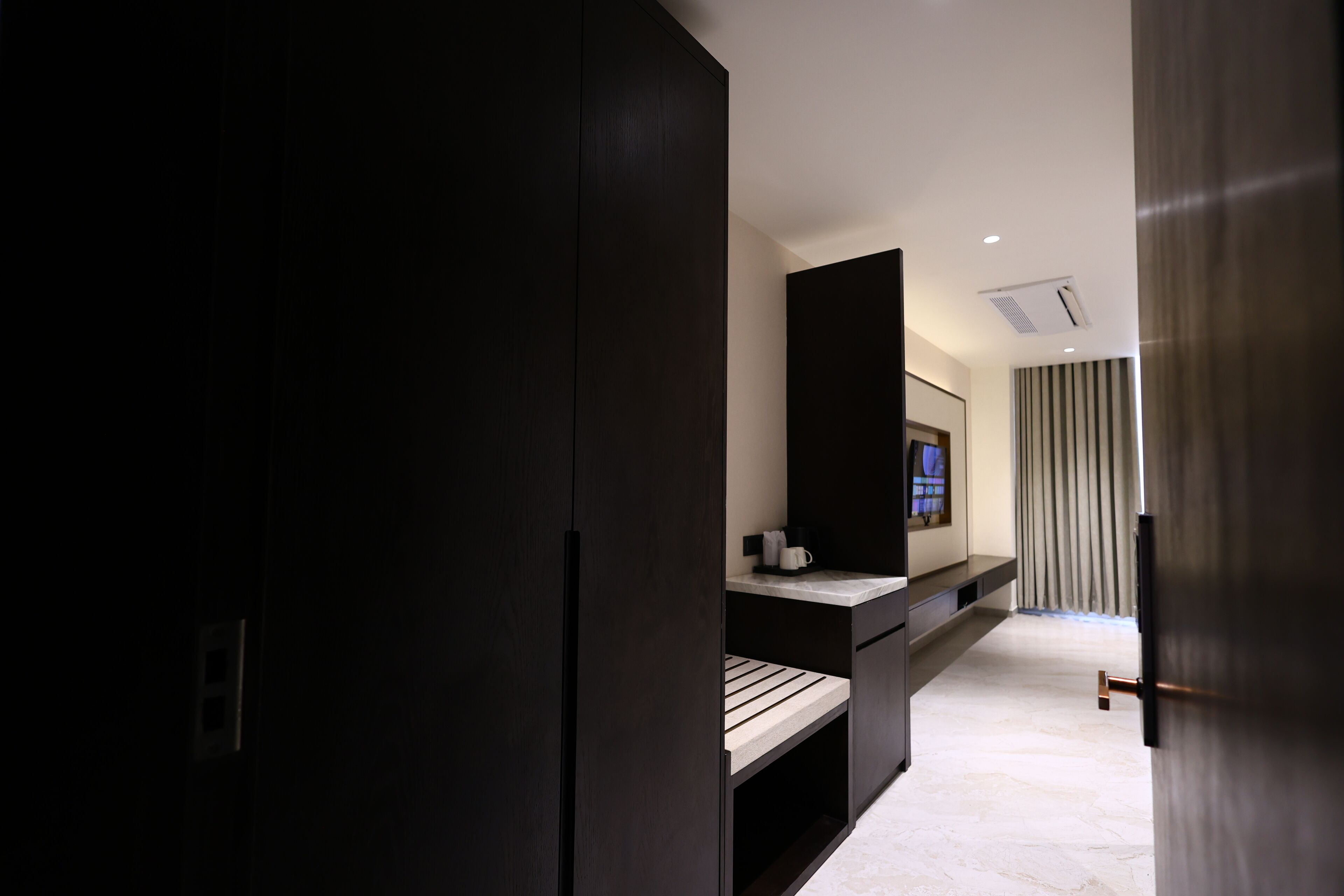 A modern hotel room with dark wood furniture, a flat-screen TV, a small seating area, and a large window with curtains.