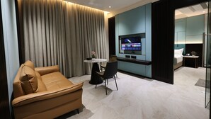A modern hotel room with a sofa, a small dining table, and a TV mounted on the wall. - ASHIRWAD CLUB RESORT (Jamnagar)