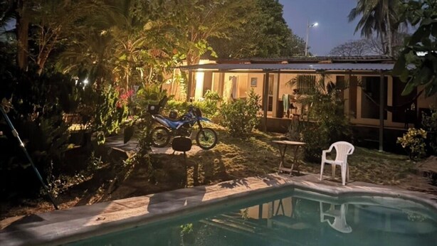 Zona de piscina nocturna con una casa al fondo, rodeada de árboles y una motocicleta.