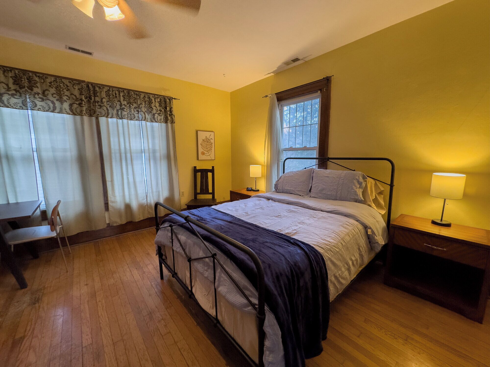 Un dormitorio con cama, lámparas de noche, una silla, una ventana con cortinas y suelo de madera.