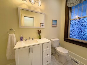 A bathroom with a white sink, mirror, and toilet. There is a window with a stained glass design, a small vase with dried flowers, and a towel hanging on a ring.