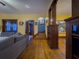 Una sala de estar con un sofá gris, piso de madera, una escalera y vista a un estanque a través de una ventana.