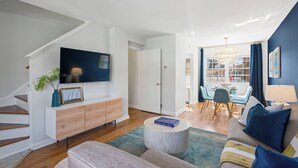A modern living room with a flat-screen TV, a wooden cabinet, a round coffee table, a sofa, and a dining area with chairs and a table. - Old Town Living- Xcape to Vibrant Old Town (Alexandria)