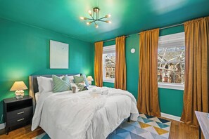 Ein Schlafzimmer mit türkisfarbenen Wänden, einem Bett mit weißer Bettwäsche, einem Nachttisch mit Lampe und Blick auf ein Gebäude durch ein Fenster mit braunen Vorhängen.