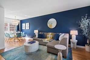 Ein modernes Wohnzimmer mit blauer Akzentwand, einem runden Spiegel, einer Couch mit Kissen, einem Couchtisch und einem Essbereich mit Stühlen und Tisch.
