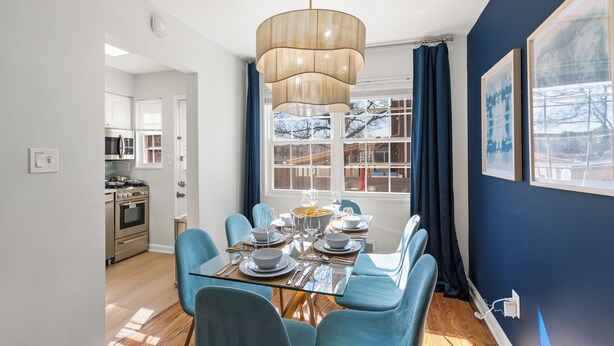 A dining area with a glass table set for six, blue chairs, a modern chandelier, and a view of the outside through large windows. - Old Town Living- Xcape to Vibrant Old Town (Alexandria)