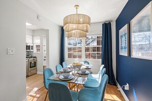 A dining area with a glass table set for six, blue chairs, a modern chandelier, and a view of the outside through large windows. - Old Town Living- Xcape to Vibrant Old Town (Alexandria)