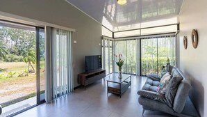 A modern living room with a sofa, coffee table, and TV, large sliding glass doors to a garden, and a ceiling light. - Villa 2 (2BR) w/Pool • Near Downtown Turrialba (Turrialba)