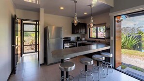 A modern kitchen with stainless steel appliances, a bar area with stools, and a large sliding glass door leading to a garden view. - Villa 2 (2BR) w/Pool • Near Downtown Turrialba (Turrialba)