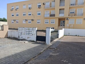 Um edifício residencial com vários andares, unidades de ar condicionado, portão fechado e entrada pavimentada.