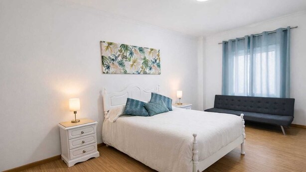 Een slaapkamer met een bed, nachtkastjes, een bankje en een raam met gordijnen.