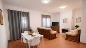 Een woonkamer met een witte tafel, twee beige fauteuils, een houten kast en een televisie.