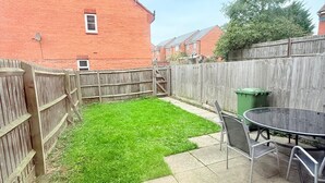 Un jardin avec une pelouse verdoyante, une clôture en bois, une maison en briques et une terrasse équipée d’une table et de chaises.