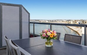 Ein Balkon mit Tisch und Stühlen, einem Blumenstrauß und Blick auf eine Uferpromenade mit Gebäuden.
