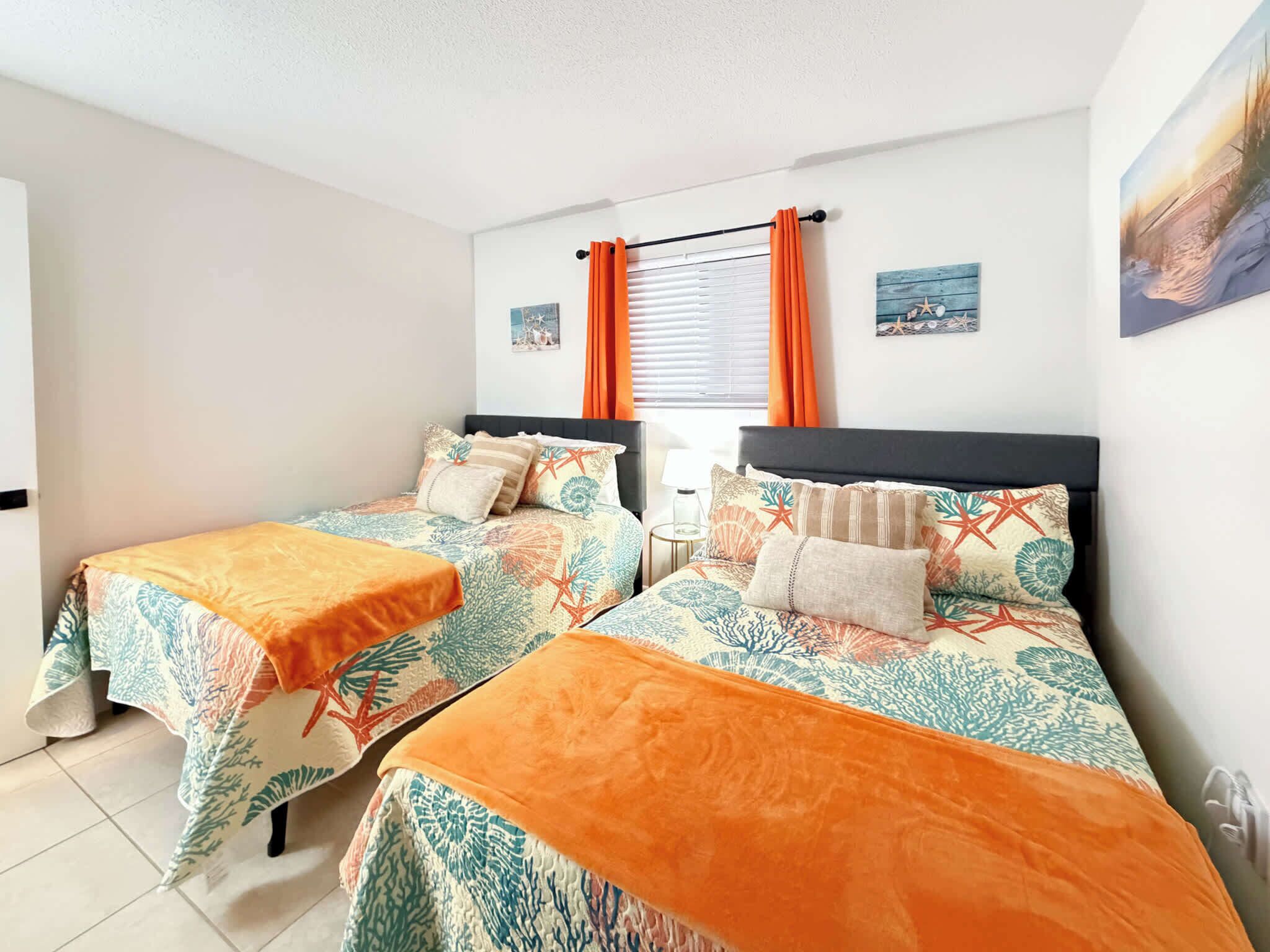 A room with two beds, each with a patterned bedspread and orange throw blankets.